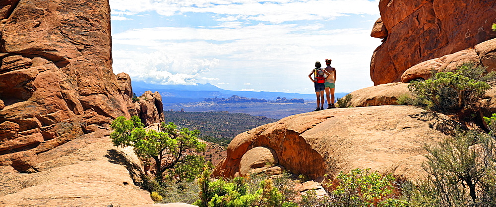 Hikers looking towards the La Sal Mountains from between red sandstone cliffs, Arches-Nationalpark, near Moab, Utah, United States