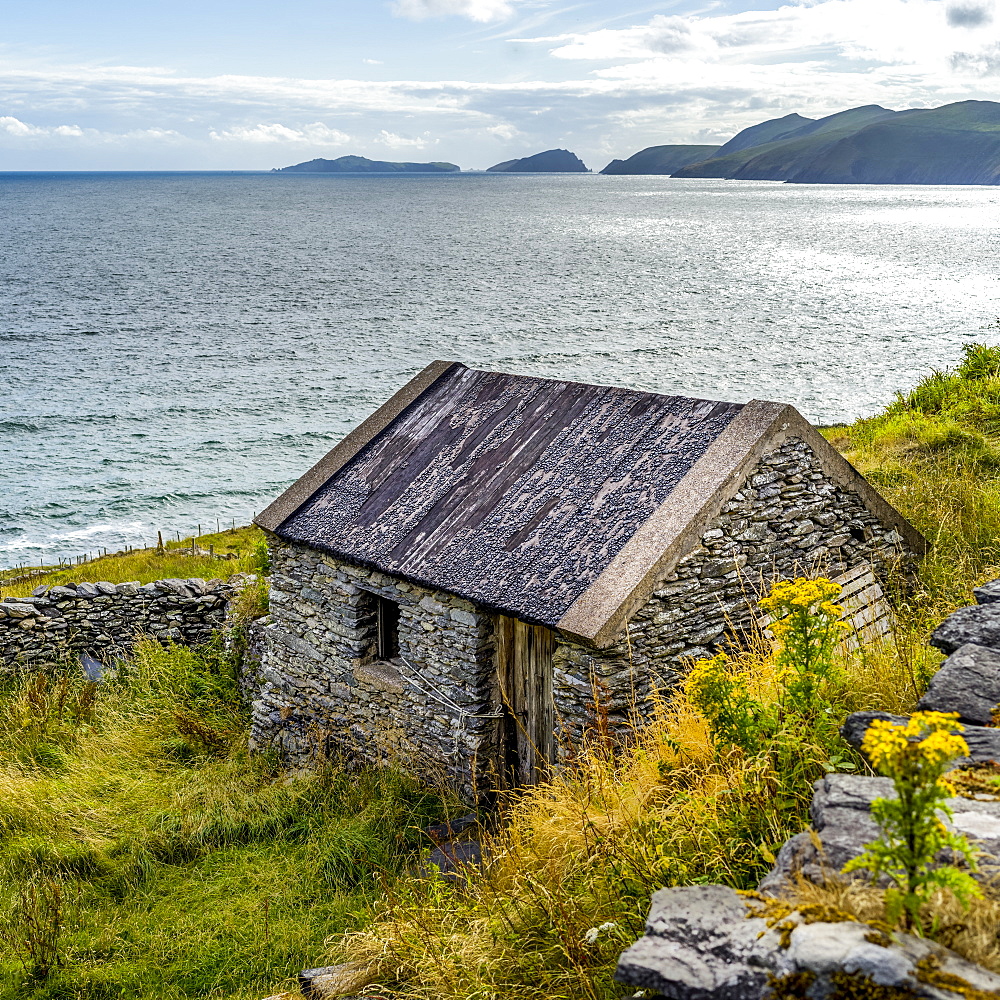 A small stone house along the coastline of County Kerry with cliffs and mountains in the distance, Ballyferriter, County Kerry, Ireland