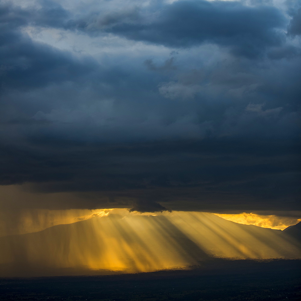 Golden sun rays and illuminated rainfall shining through the dark, ominous clouds with mountains in the distance, Volcanoes National Park, Northern Province, Rwanda