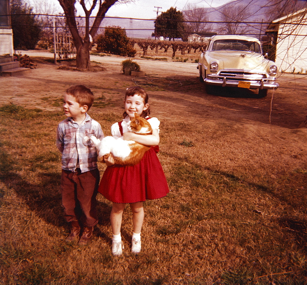 Caucasian brother and sister standing in yard holding cat