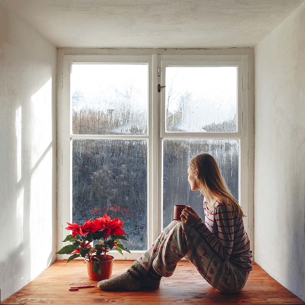 Caucasian woman sitting in window sill drinking coffee in pajamas