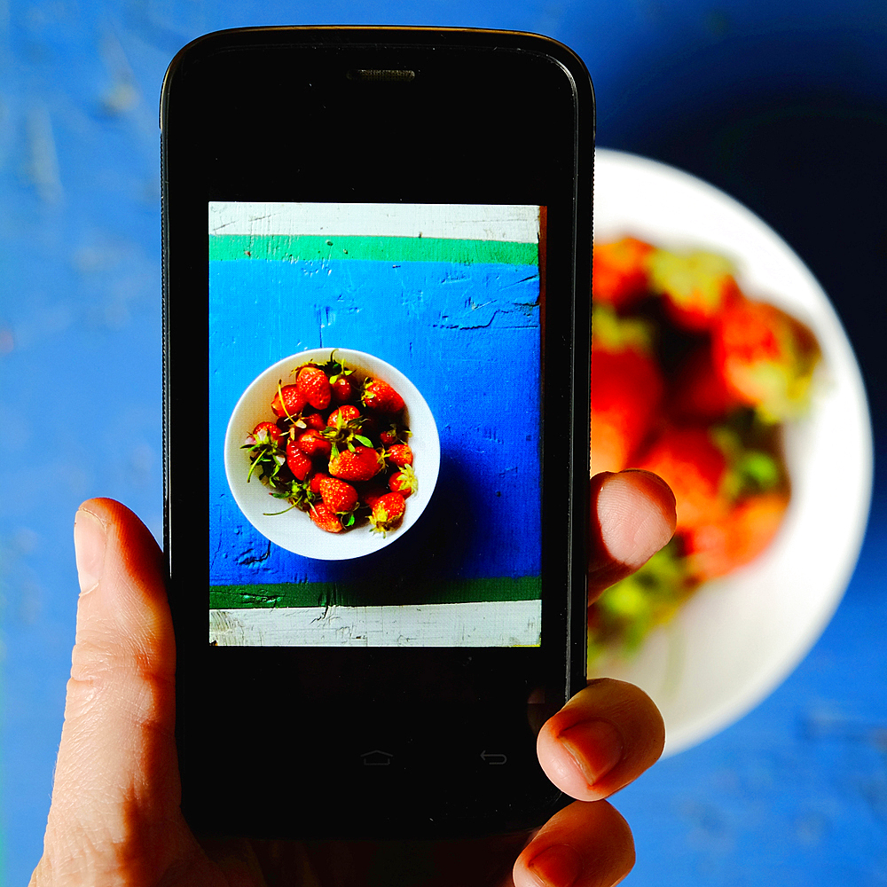 Hand of Caucasian woman photographing bowl of strawberries