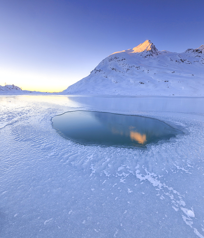 The sun illuminating a peak surrounding the frozen Lago Bianco by Bernina, reflecting in a pool in the center of the lake, Bernina Pass, Graubunden, Swiss Alps, Switzerland, Europe