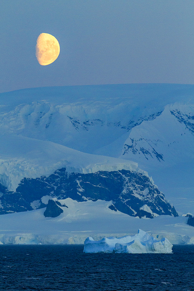 View of the nearly full moon rising over snow-covered mountains on the Antarctic Peninsula, Antarctica.