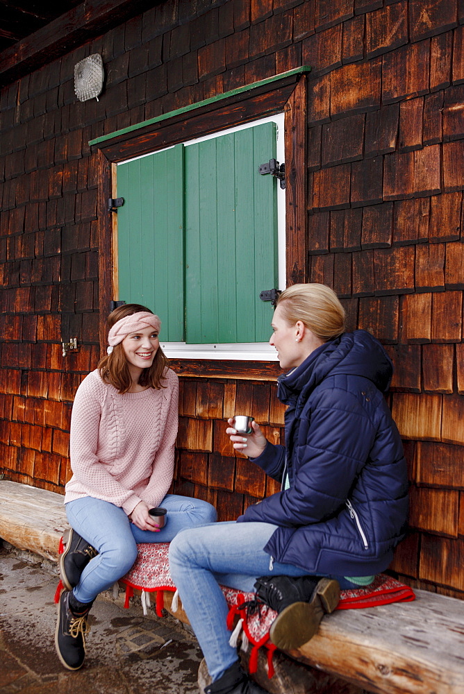 Two young women on a bench having a hot beverage, Spitzingsee, Upper Bavaria, Germany