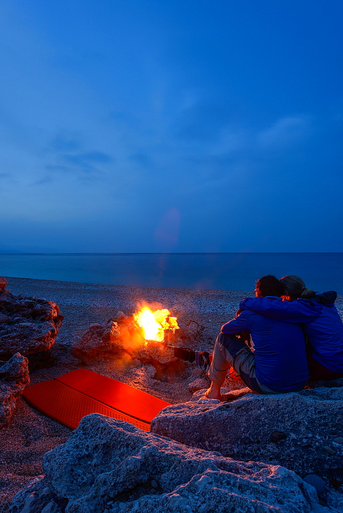 Young woman and young man sitting on rocks near the camp fire and camping out on the pebble beach of the bay Cala Sisine, Golfo di Orosei, Selvaggio Blu, Sardinia, Italy, Europe