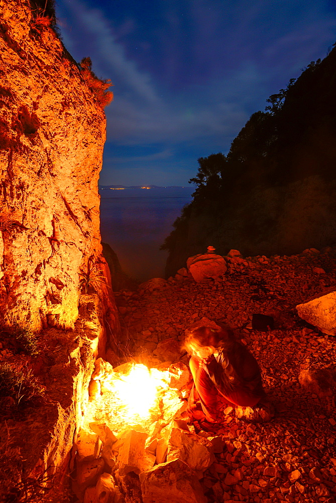 A young woman sitting at the camp fire above the sea near Bacu Mudaloru in the mountainous coast landscape, Golfo di Orosei, Selvaggio Blu, Sardinia, Italy, Europe