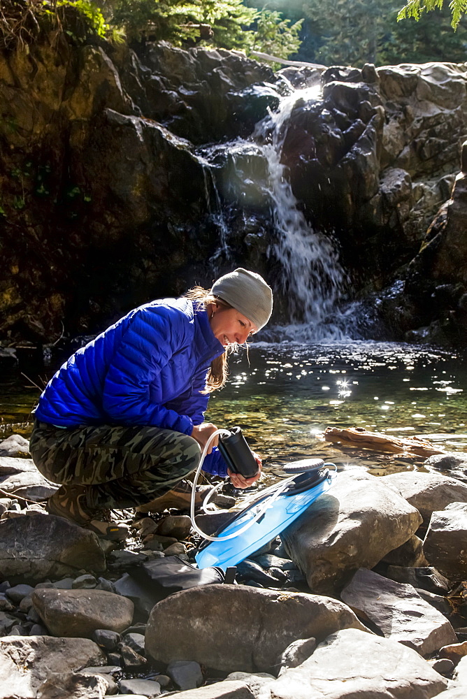 Female backpacker filtering water, backlit by morning sunshine on the High Divide Trail, with waterfall in background, Olympic National Park, Olympic Mountains, Washington, United States of America