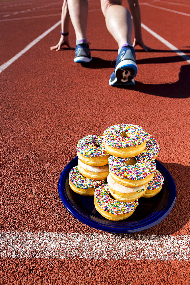 A plate full of candy covered donuts on a running track with a female runner in a lane in a starter stance, Calgary, Alberta, Canada