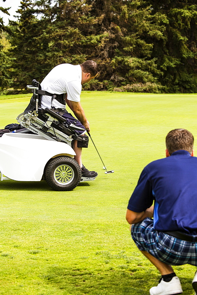 An able bodied golfer teams up with a disabled golfer using a specialized powered golf wheelchair and helps him to line up his putt on a golf green at a golf course, Edmonton, Alberta, Canada