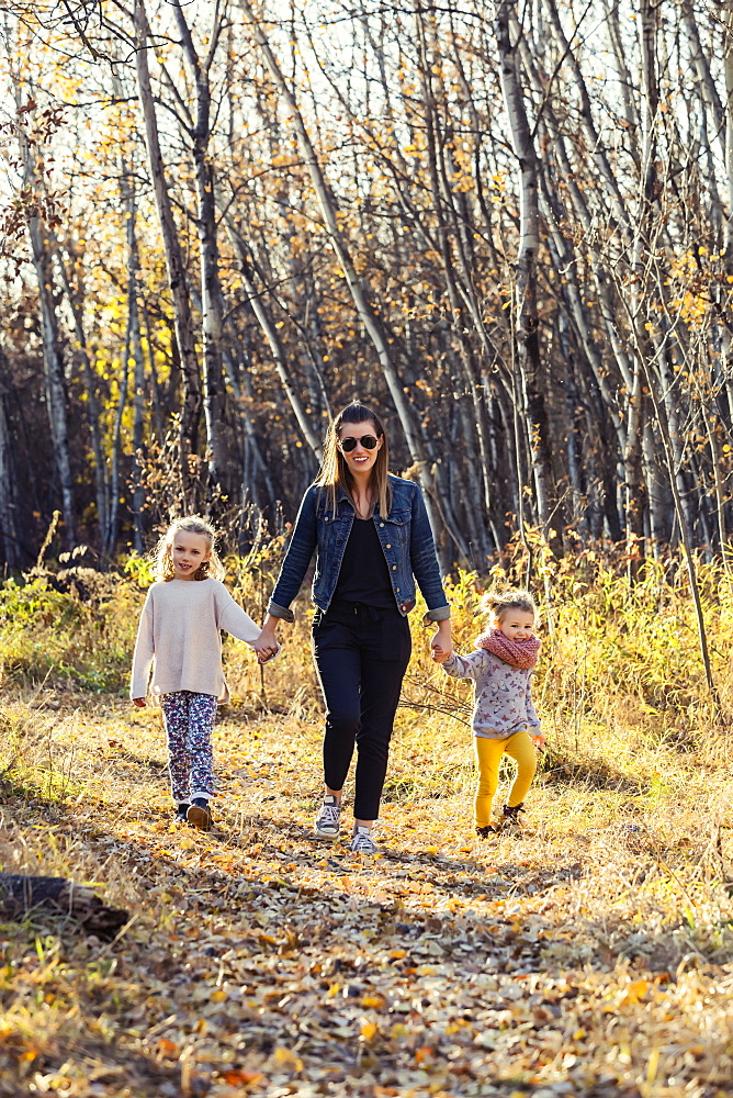 A mother and her two little daughters walking through the woods and holding hands in a city park on a warm fall evening, Edmonton, Alberta, Canada