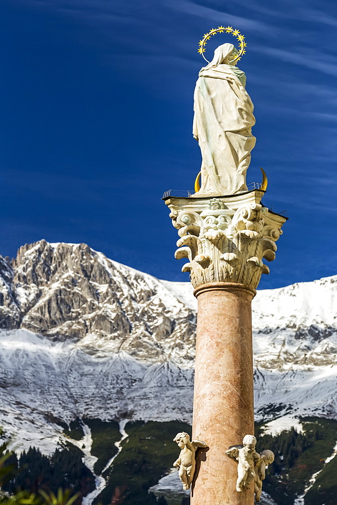 Stone statue with halo on top of column with snow-covered mountain range and blue sky in the background, Innsbruck, Tyrol, Austria