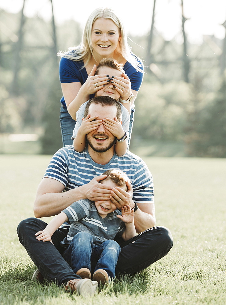 Portrait of a family with young children in a park, standing in a row covering each other's eyes, Edmonton, Alberta, Canada