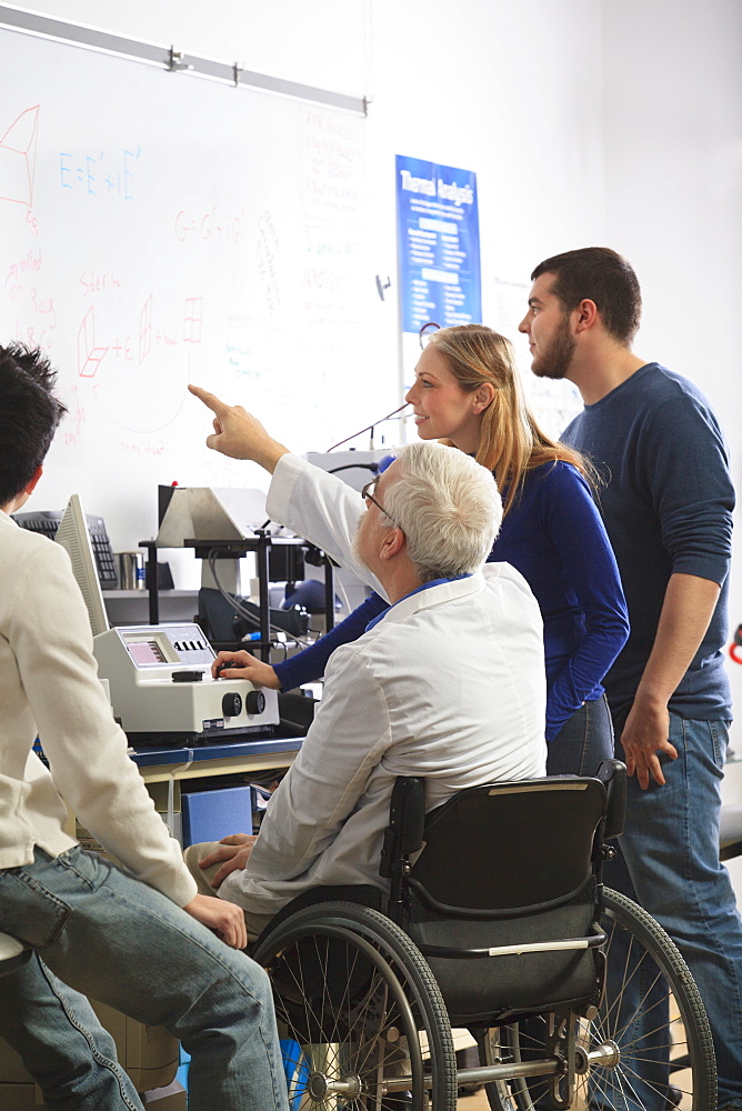 Professor with muscular dystrophy and engineering students reviewing analyzer results on white board in a laboratory