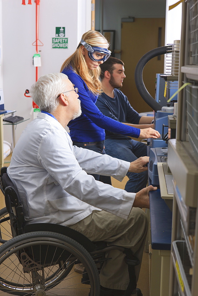 Professor with muscular dystrophy advising engineering students in chemistry laboratory working on thermogravimetric analyzers