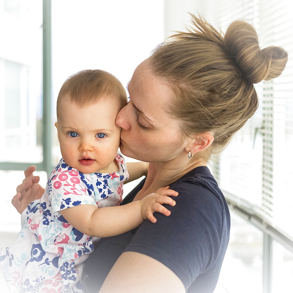 Portrait of infant baby girl with mother at home, mother kissing baby on the cheek; Vancouver, British Columbia, Canada