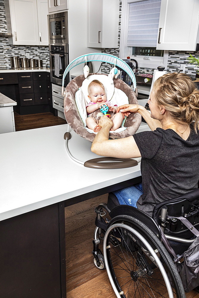 A paraplegic mother in a wheelchair playing with her baby in a baby seat, while in her kitchen; Edmonton, Alberta, Canada