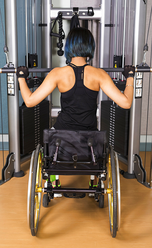 A view from behind of a paraplegic woman working out using a lat pull down machine in a fitness facility; Sherwood Park, Alberta, Canada