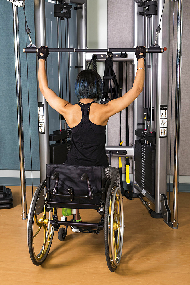 A view from behind of a paraplegic woman working out using a lat pull down machine in a fitness facility; Sherwood Park, Alberta, Canada