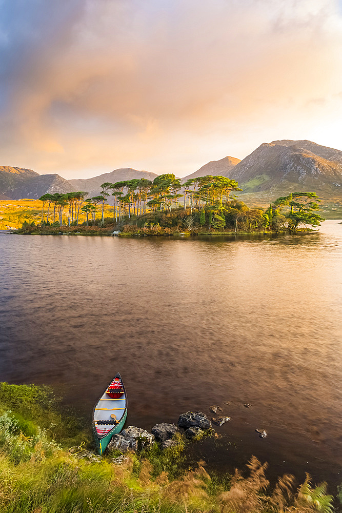 Canoe on the bank of a lake pointing towards an island with pine trees with an epic sunrise in the background; Connemara, County Galway, Ireland