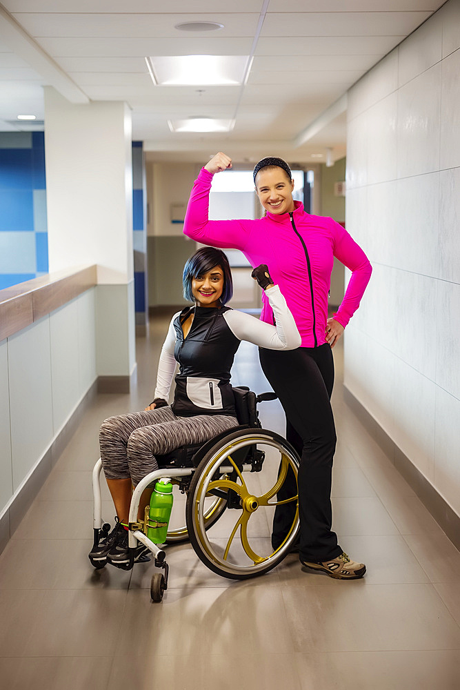 A paraplegic woman and her trainer show off their muscles while posing for the camera in a hallway in a recreational facility: Sherwood Park, Alberta, Canada