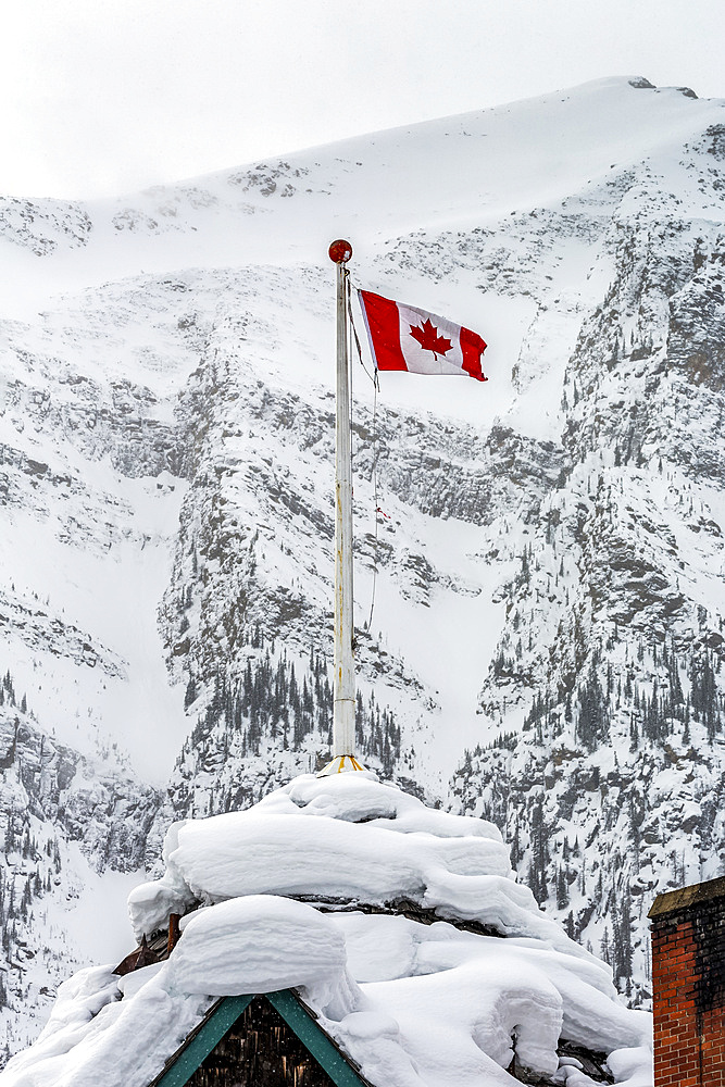 Close up of snow-covered building peak with a Canadian flag and a snow-covered mountain in the background, Banff National Park; Lake Louise, Alberta, Canada