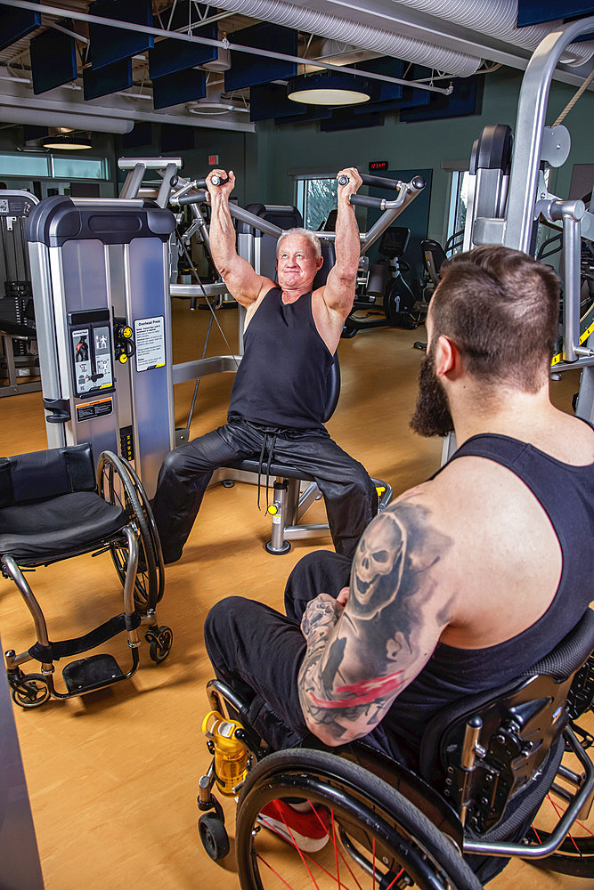 One paraplegic man working out using an overhead press in fitness facility while his disabled friend watches; Sherwood Park, Alberta, Canada