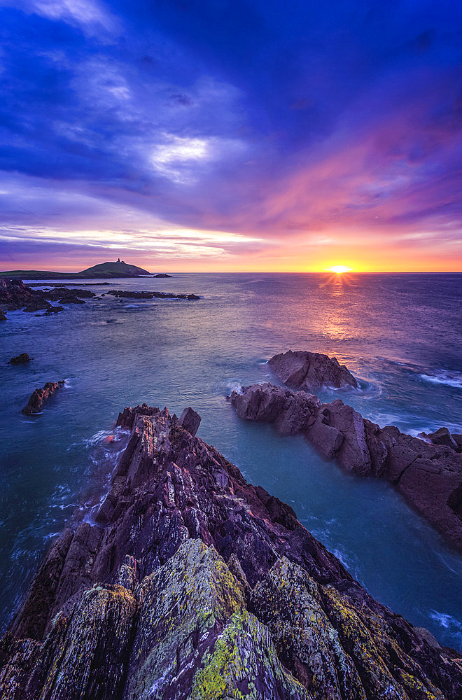 Sunrise along the Irish coast with rock formations in the foreground and Ballycotton lighthouse on the horizon; Ballycotton, County Cork, Ireland