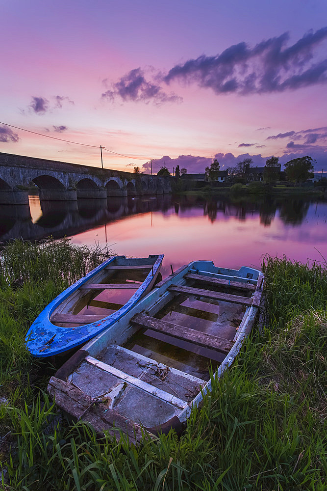 Two old wooden boats on the banks of the Shannon River at sunset with a stone bridge in the background; Montpellier, County Limerick, Ireland