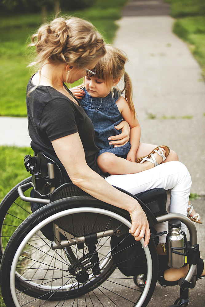 A paraplegic mom holding her little girl in her lap while taking her for a ride down the street in her wheelchair on a warm summer afternoon: Edmonton, Alberta, Canada.