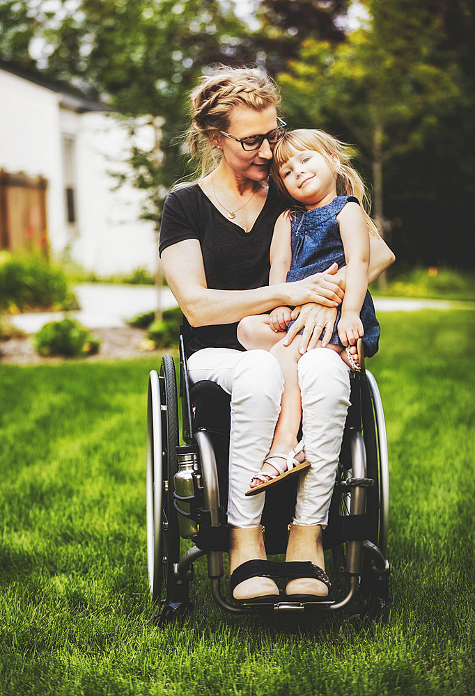 A paraplegic mom holding her little girl in her lap while sitting in her wheelchair in her front yard on a warm summer afternoon: Edmonton, Alberta, Canada.