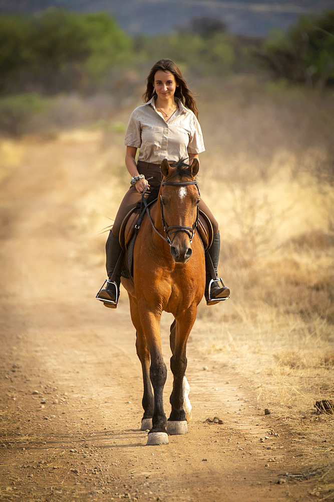 Woman riding horse (Equus ferus caballus) on a dirt road through the bush on the savanna looking at the camera and smiling at the Gabus Game Ranch; Otavi, Otjozondjupa, Namibia