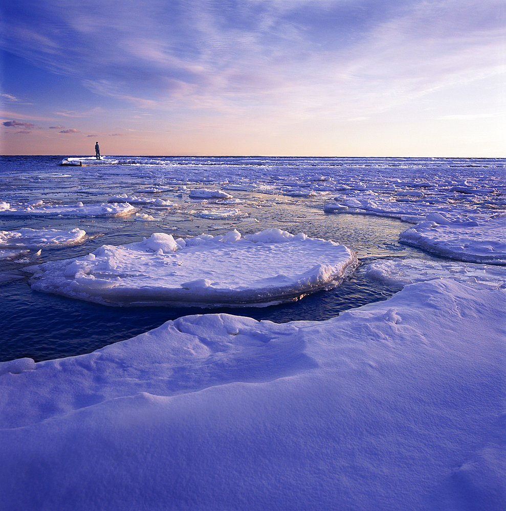 Person stands on the edge of an ice formation in the ocean and looks out to the vast seascape and horizon