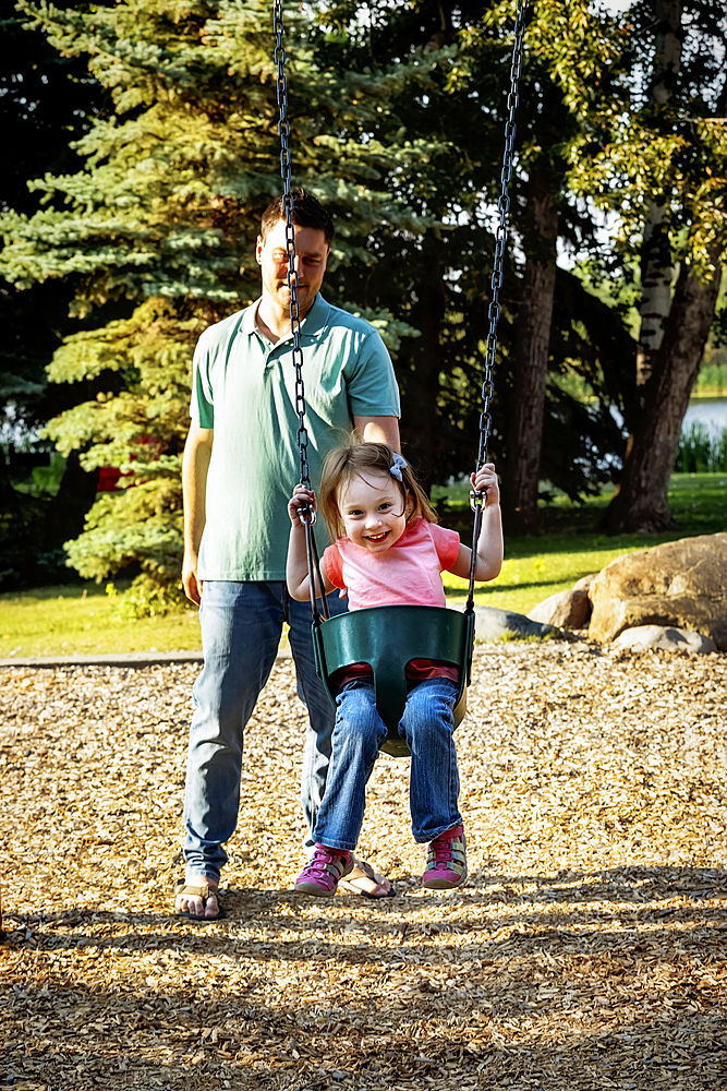 Father spending quality time with his young daughter and pushing her on a swing in a city park, during a warm fall afternoon, Leduc, Alberta, Canada