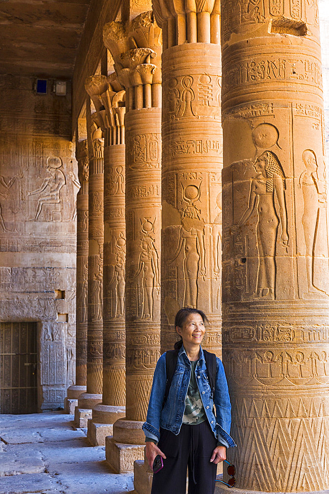 A female Chinese tourist stands among a row of columns with ancient Egyptian hieroglyphic details at the Philae Temple, Aswan, Egypt, Africa