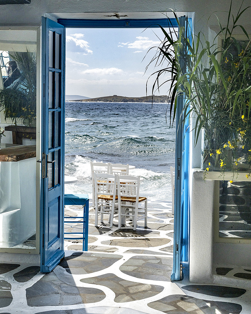 Blue doors wide open to the ocean air and sunlight, with the surf washing onto a patio with chairs, Mykonos, Greece