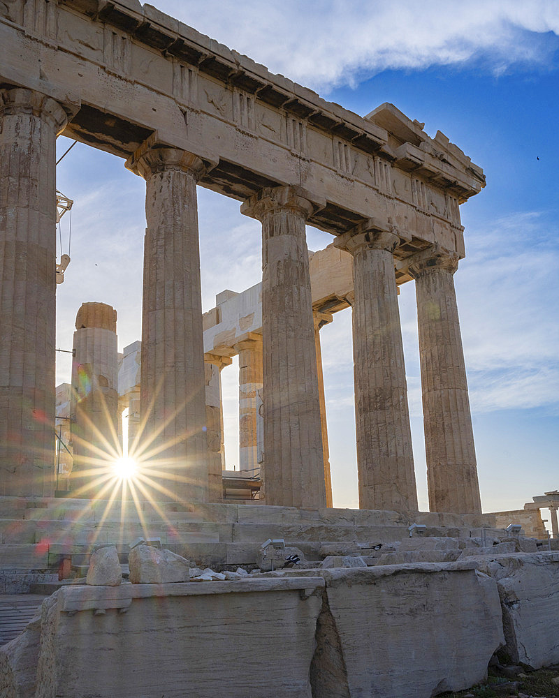 Close-up view of the columns of The Parthenon against a blue, cloudy sky with a sunburst at twilight on the Acropolis of Athens, an ancient citadel located on a rocky outcrop above the City of Athens, Athens, Greece