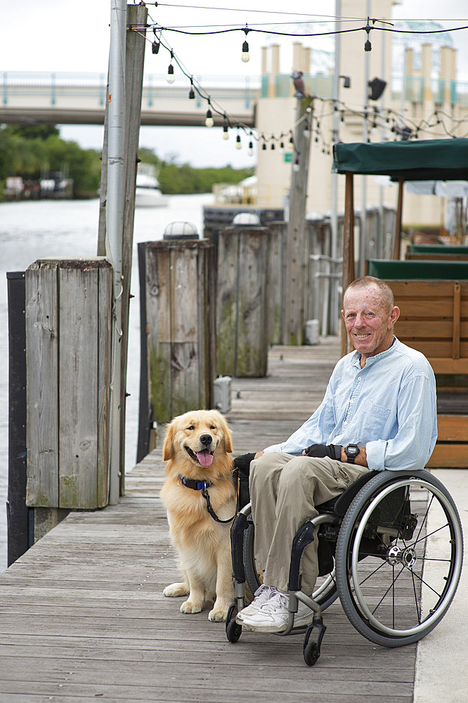 Informal outdoor portrait of a man in a wheelchair with his service dog sitting on a boardwalk along a waterfront, Boynton Beach, Florida, United States of America