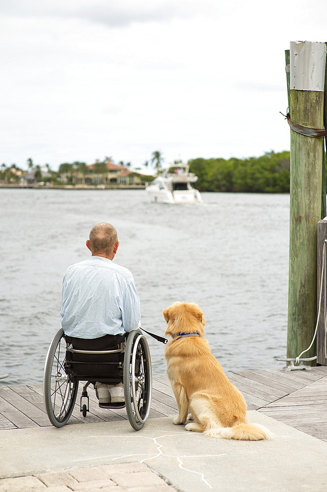 Man in a wheelchair with his service dog sitting on a boardwalk at the water's edge enjoying a waterfront view, Boynton Beach, Florida, United States of America