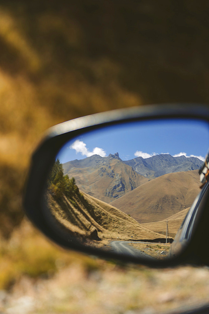 Vehicle rear view mirror reflects a mountain range on a sunny day in Kazbegi National Park, Georgia