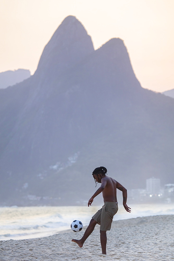 A young black Brazilian playing football on Ipanema beach with the Dois Irmaos mountains in the distance, Rio de Janeiro, Brazil, South America