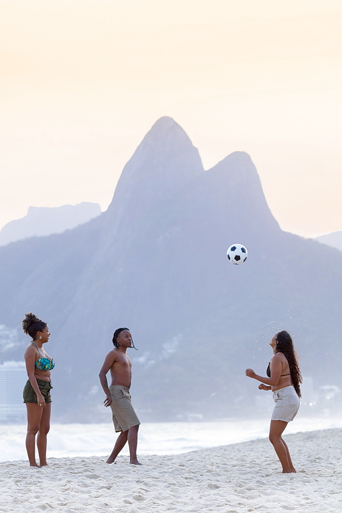 A young black Brazilian playing football with two female friends on Ipanema beach with the Dois Irmaos mountains in the distance, Rio de Janeiro, Brazil, South America