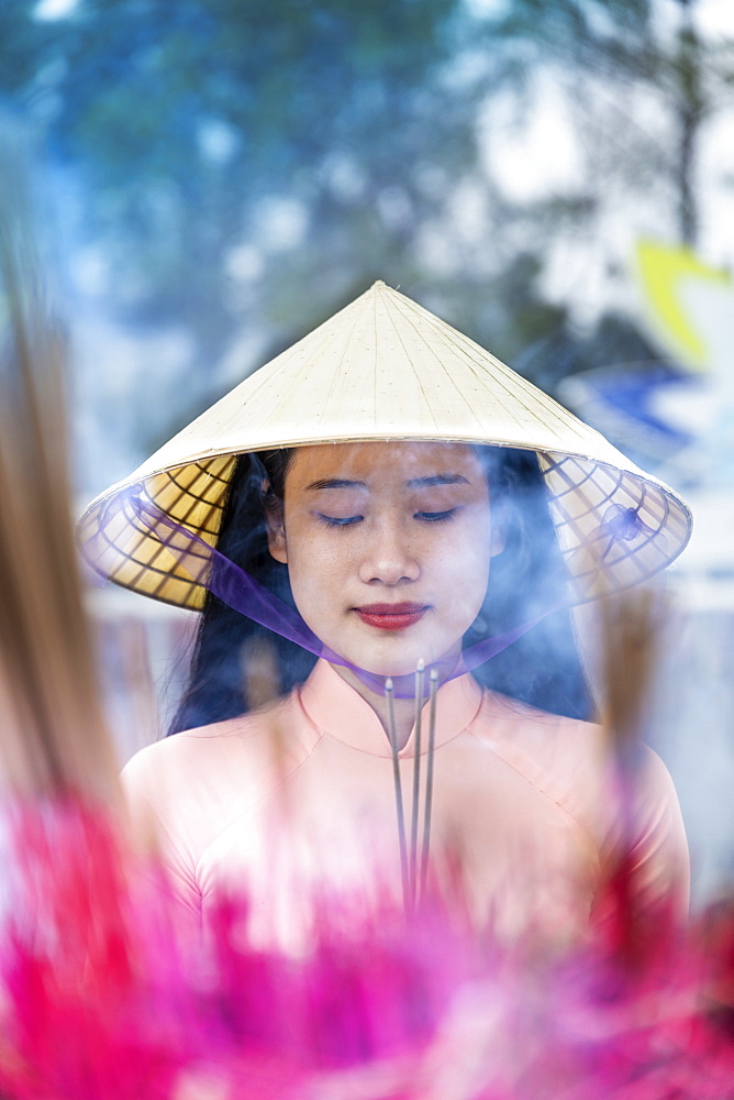 A young Vietnamese woman in a conical hat making incense offerings at a Buddhist temple, Hue, Vietnam, Indochina, Southeast Asia, Asia