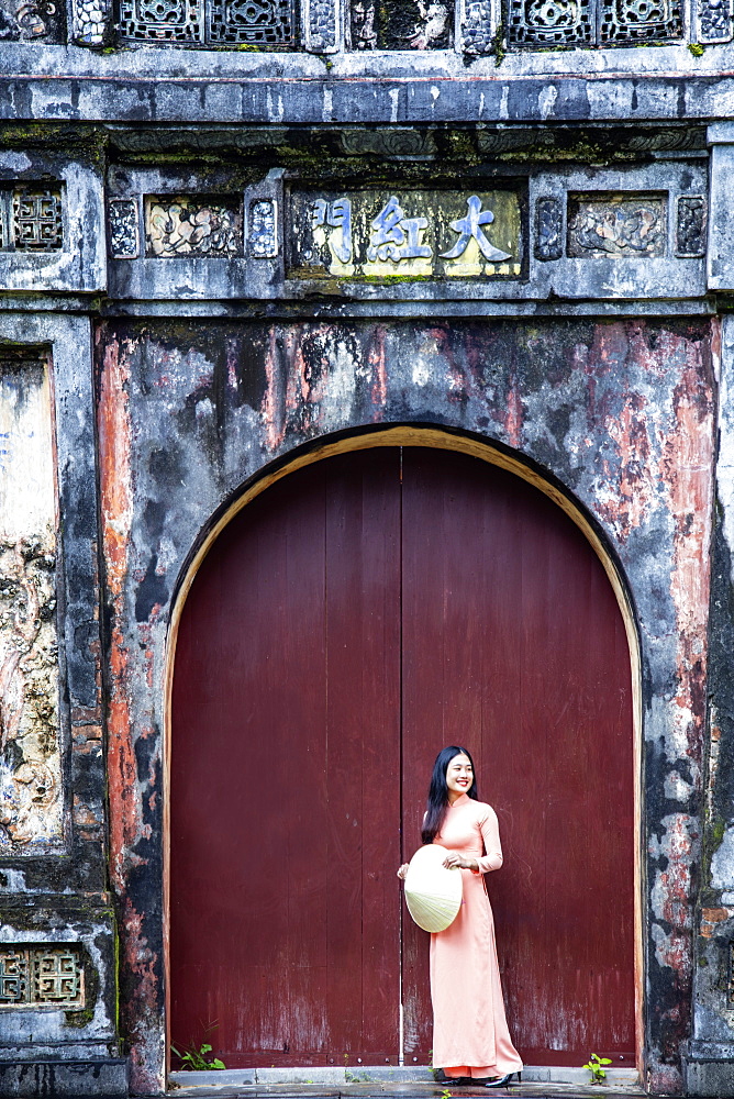 A young Vietnamese woman in a traditional Ao Dai dress and hat standing at the gateway to the Imperial Purple City, Hue, Vietnam, Indochina, Southeast Asia, Asia