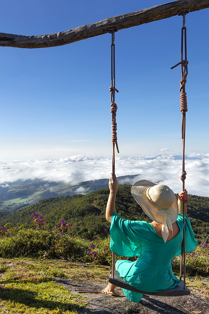 A young woman using a swing over a view of rainforest-covered mountains in the Ibitipoca Reserve, Minas Gerais, Brazil, South America