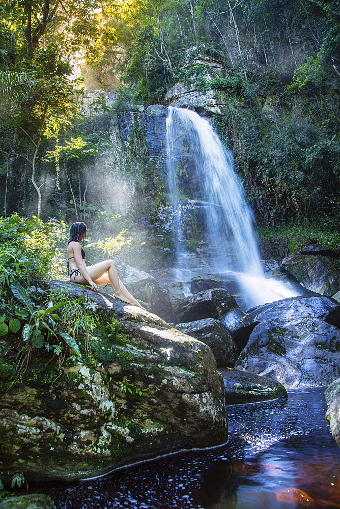 A beautiful young woman sitting in a shaft of sunlight in front of a pristine waterfall in the rainforest, Brazil, South America