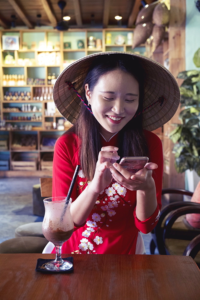 A young Asian woman in a red Ao Dai dress and conical hat smiling and using a mobile phone in a cafe setting, Hoi An, Vietnam, Indochina, Southeast Asia, Asia