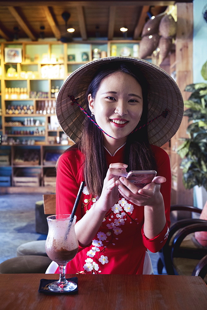 A young Asian woman in a red Ao Dai dress and conical hat smiling and using a mobile phone in a cafe setting, Hoi An, Vietnam, Indochina, Southeast Asia, Asia