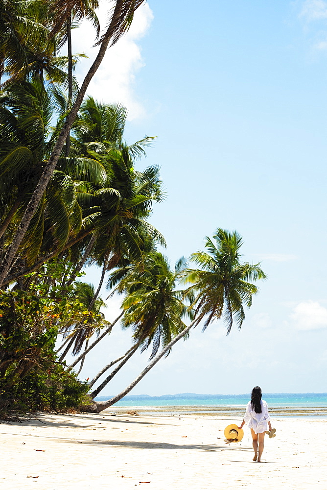 A young woman with brown hair wearing a white beach shirt and holding a hat walking along a tropical beach, Brazil, South America