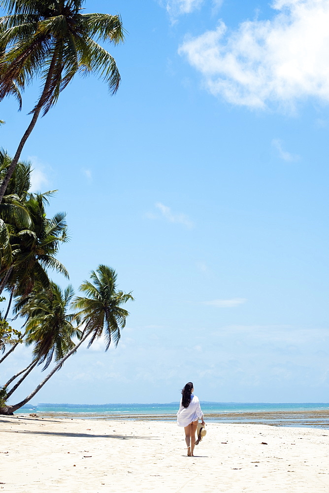 A young woman with brown hair wearing a white beach shirt and holding a hat walking along a tropical beach, Brazil, South America